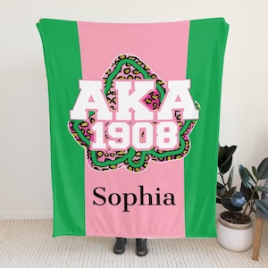 Op de afbeelding: Een groene, roze en witte deken met de tekst "AKA 1908 Sophia". De deken heeft een roze middenstuk met een luipaardprint en groene strepen aan weerszijden. De tekst "AKA 1908" staat in witte blokletters.