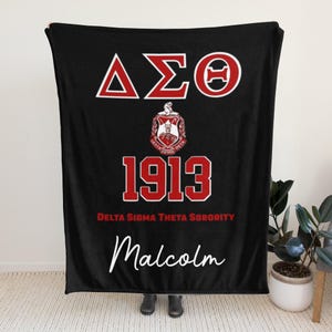 Op de afbeelding: Zwarte deken met rode en witte Griekse letters, het nummer 1913 en de woorden "Delta Sigma Theta Sorority". De deken heeft ook de naam "Malcolm" in witte letters. Een decoratief item voor een sorority-lid.
