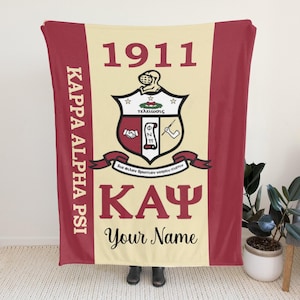 Op de afbeelding: Een bordeauxrode en crèmekleurige deken met de tekst "KAPPA ALPHA PSI" verticaal aan de linkerkant. Het midden toont "1911" boven een wapen en "KAY" met "Your Name" eronder. De deken heeft bordeauxrode randen.