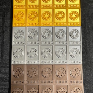 Könnte beinhalten: Reihen rechteckiger Spielsteine in Gold, Silber und Bronze. Jedes Teil zeigt ein kreisförmiges Emblem und die Zahl 7. Die goldenen Teile haben ein Sternmuster, während die silbernen und bronzenen Teile ein anderes Emblem haben.