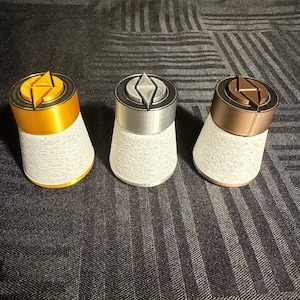 May include: Three decorative bottle stoppers with geometric designs. One is gold, another silver, and the third is bronze. Each has a white, textured base. The stoppers are arranged on a dark, patterned surface.