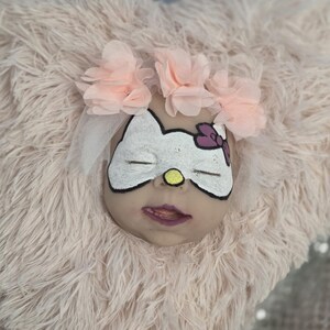 Puede incluir: Una cabeza de muñeca con una cara de Hello Kitty pintada, ojos blancos y una nariz amarilla. La cabeza está adornada con una corona de flores rosas y un velo transparente. La cabeza descansa sobre una superficie rosa claro y esponjosa.