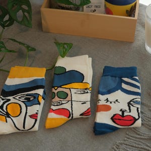 Pode incluir: Três pares de meias brancas com desenhos de rostos abstratos. Cada meia apresenta um rosto diferente com linhas arrojadas e detalhes coloridos em amarelo, azul, vermelho e laranja. As meias são exibidas em uma superfície cinza.