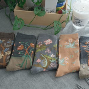 Può includere: Cinque paia di calzini fantasia con disegni floreali e botanici. I calzini presentano vari schemi di colori, tra cui marrone, grigio e pesca, con illustrazioni di fiori, piante e uccelli. I calzini sono disposti su una superficie grigia.