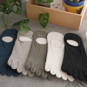 May include: Five pairs of toe socks in navy blue, light grey, olive green, white, and black. Each sock has individual toe compartments and a foot cutout. The socks are displayed on a grey surface. A candle and plant are in the background.