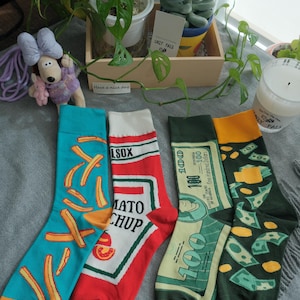 May include: Four pairs of novelty socks with distinctive designs. One pair features fries, another ketchup, a third a hundred-dollar bill, and the last with money falling. The socks are in various colours, including blue, red, green, and yellow.