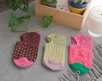May include: Three pairs of no-show socks in various colors and patterns are displayed on a gray surface. One sock is maroon with yellow polka dots, another is light yellow with a ribbed design, and the third is pink with a red pattern.