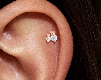 Puede incluir: Primer plano de una oreja adornada con dos pendientes pequeños de color dorado. Cada pendiente presenta una brillante gema redonda y transparente. Los pendientes tienen un diseño delicado y minimalista.