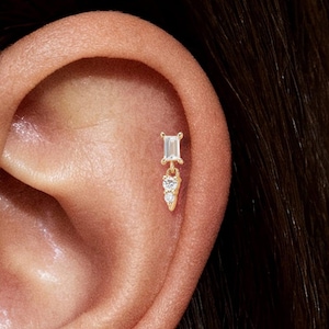 May include: A gold-colored earring is shown in an ear piercing. The earring has a rectangular top with a small dangling design below it. The dangling design has two small, round, clear stones.