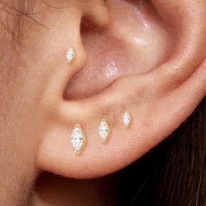 May include: Close-up of an ear adorned with multiple gold earrings. Each earring features a marquise-cut, clear gemstone. The earrings are of varying sizes and are placed along the ear's cartilage and lobe.