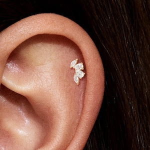 May include: A close-up of an ear with a small, gold earring. The earring has three small, clear gemstones in a floral arrangement. The ear is light-toned with visible skin texture and dark hair visible in the background.