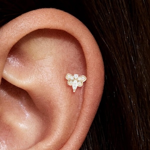 May include: A gold-colored ear piercing featuring a bee design. The bee has a body made of a pear-shaped gem and a head and wings adorned with smaller, clear gems. The piercing is set against the skin of an ear.