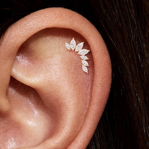 May include: Close-up of an ear featuring a cluster of small, sparkling, marquise-cut gemstones arranged in a curved design. The gemstones are set in a light-coloured metal, creating a delicate and elegant look. The jewelry is likely an ear piercing.