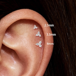 May include: An ear with three small, floral-shaped stud earrings, each set with clear gemstones. The image shows the earrings in sizes 2.5mm, 3.5mm, and 4mm. The earrings are small and sparkly, with a delicate design.