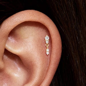 May include: Close-up of a person's ear adorned with two gold earrings. Each earring features a small, sparkling diamond at the top, with two smaller diamonds dangling below. The earrings are set against the backdrop of the ear's natural skin tone.