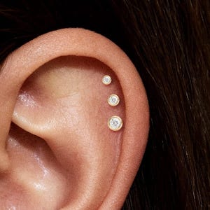 May include: Close-up of an ear adorned with multiple small, gold-colored stud earrings. Each earring features a clear, round gemstone set within a delicate bezel. The earrings are positioned along the ear's cartilage and lobe.