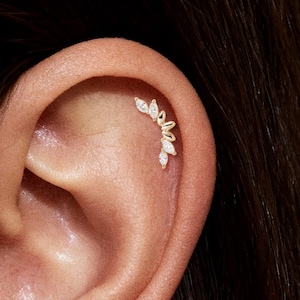 Op de afbeelding: Een close-up van een oor met een gouden helix piercing. De piercing heeft een gebogen ontwerp met verschillende heldere, druppelvormige edelstenen. Het oor is licht getint en de achtergrond is donker.