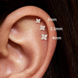 May include: A close-up of an ear with multiple small, gold-colored flower-shaped earrings. The earrings are adorned with small, clear gemstones. Text indicates sizes: 3mm, 3.5mm, and 4mm. The earrings are displayed on the ear.