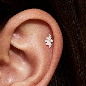 May include: A small, flower-shaped earring with multiple clear gemstones is shown in the upper ear. The earring is set in a silver or white metal. The ear is against a dark background.
