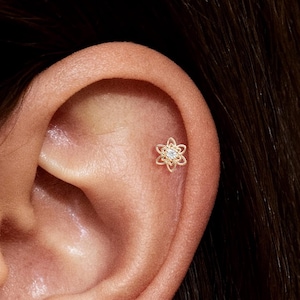 May include: A gold-toned stud earring in the shape of a six-petaled flower with a small, clear, round-cut gemstone set in the center. The earring is positioned in the upper cartilage area of the ear, featuring a delicate, open-wire metal design.