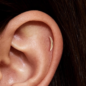 Könnte beinhalten: Nahaufnahme eines Ohrs mit einem kleinen, gebogenen goldenen Ohrring. Der Ohrring hat eine Halbmondform und sitzt am oberen Knorpel. Der Hautton ist hell, und der Hintergrund ist unscharf.