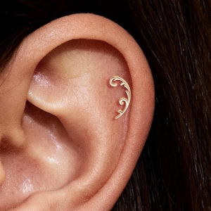 May include: Close-up of an ear adorned with two delicate, gold-colored earrings. One earring is placed on the upper ear, and the other is on the lower ear. Both earrings feature a swirling, leaf-like design, adding a touch of elegance.