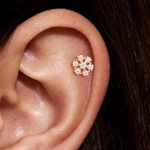 Pode incluir: Close-up de uma orelha adornada com dois brincos de botão dourados em forma de flor. Cada brinco apresenta um grupo de pequenas pedras preciosas transparentes, criando um design floral brilhante. Os brincos estão colocados na pele.
