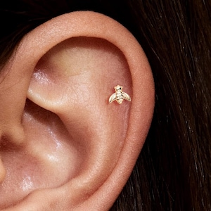 May include: A close-up of an ear with a small, gold-colored bee stud earring. The bee has detailed wings and body, and is positioned on the upper ear cartilage. The skin tone is light, and the earring is the focal point.