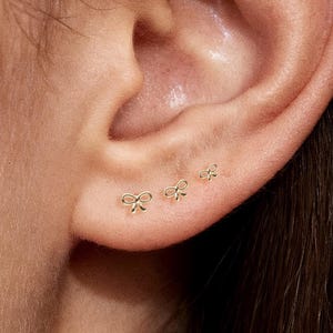 Peut inclure: Gros plan sur une oreille avec trois petites boucles d'oreilles dorées en forme de nœud dans le lobe et une dans le cartilage supérieur. Les boucles d'oreilles sont délicates et minimalistes.