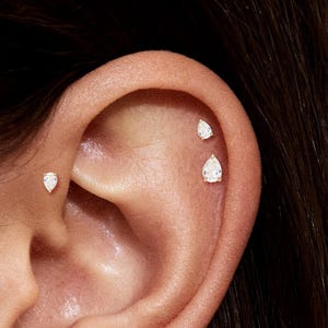 Może przedstawiać: Zbliżenie ucha z trzema małymi, błyszczącymi kolczykami w kształcie łezki. Kolczyki osadzone są w jasnym metalu i umieszczone na górnej części ucha i chrząstce. Odcień skóry jest jasny.