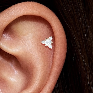 Op de afbeelding: Een close-up van een oor met een kleine, driehoekige oorbel. De oorbel is zilverkleurig en heeft drie kleine, heldere edelstenen. Het oor is licht getint met zichtbare huidtextuur.