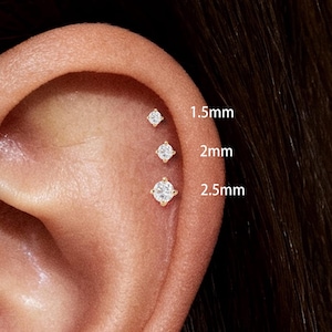 May include: Close-up of an ear with multiple small, sparkling diamond stud earrings. The earrings are labeled as 1.5mm, 2mm, and 2.5mm. The earrings are set in gold. The ear is against a neutral background.