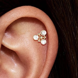 以下が含まれることがあります： 3つの透明な丸い宝石がクラスターデザインになった、ゴールドトーンのヘリックスピアス。ピアスは耳に装着され、その配置を示しています。デザインは繊細でエレガントで、ヘリックスピアスに適しています。