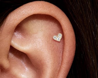 Puede incluir: Un pequeño pendiente en forma de corazón con múltiples piedras pequeñas y transparentes engastadas en un metal dorado. El pendiente está colocado en el cartílago superior de la oreja. La imagen muestra un primer plano de la oreja y el pendiente.