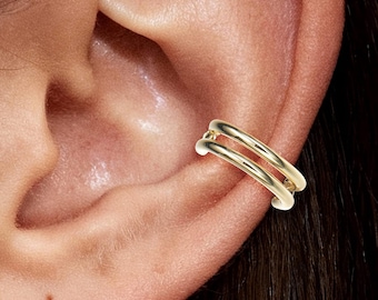 May include: A gold-colored ear cuff featuring a double-band design. The cuff is shown on the ear, highlighting its minimalist and modern aesthetic. The jewelry piece is designed to be worn without piercing.