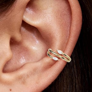 以下が含まれることがあります： 2つの白い涙型の宝石が付いたゴールドのイヤーカフ。 イヤーカフは耳の軟骨に巻き付き、モダンでミニマルなジュエリーデザインを作り出します。 宝石は繊細で磨かれたゴールドセッティングにセットされています。