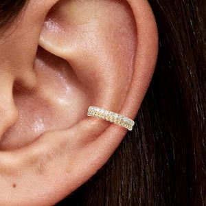 Op de afbeelding: Een goudkleurige oormanchet met twee rijen kleine, heldere edelstenen. De manchet is ontworpen om rond het kraakbeen van het oor te wikkelen. De edelstenen zijn dicht op elkaar geplaatst, wat een sprankelend effect creëert.