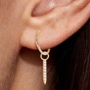 以下が含まれることがあります： 小さな透明な宝石がちりばめられた、ぶら下がっている垂直のバーが付いたゴールドのフープイヤリング。 イヤリングはクローズアップビューで表示され、耳を背景にジュエリーの詳細を強調しています。