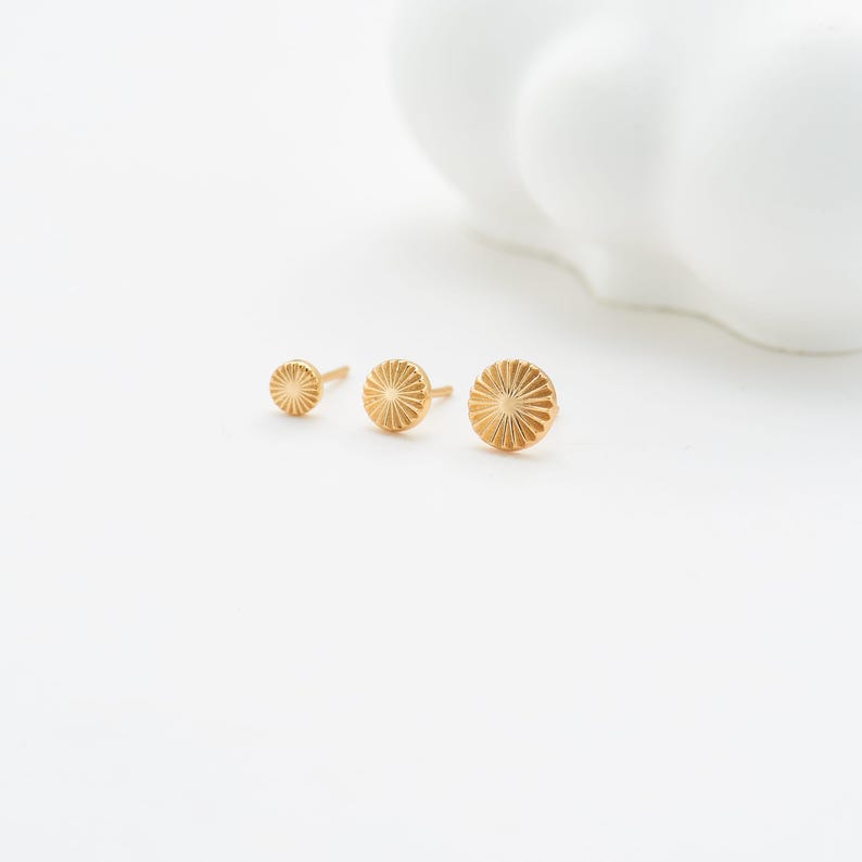 May include: Three gold-colored stud earrings in varying sizes. Each earring features a sunburst design with radiating lines. The earrings are displayed on a white surface, with a blurred white object in the background.