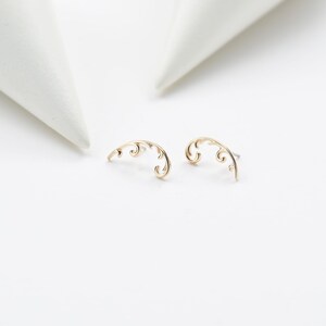 May include: A pair of gold-colored, curved earrings with an ornate, swirling design. The earrings are set against a white background, with two white, cone-shaped objects in the background. The earrings are delicate and elegant.