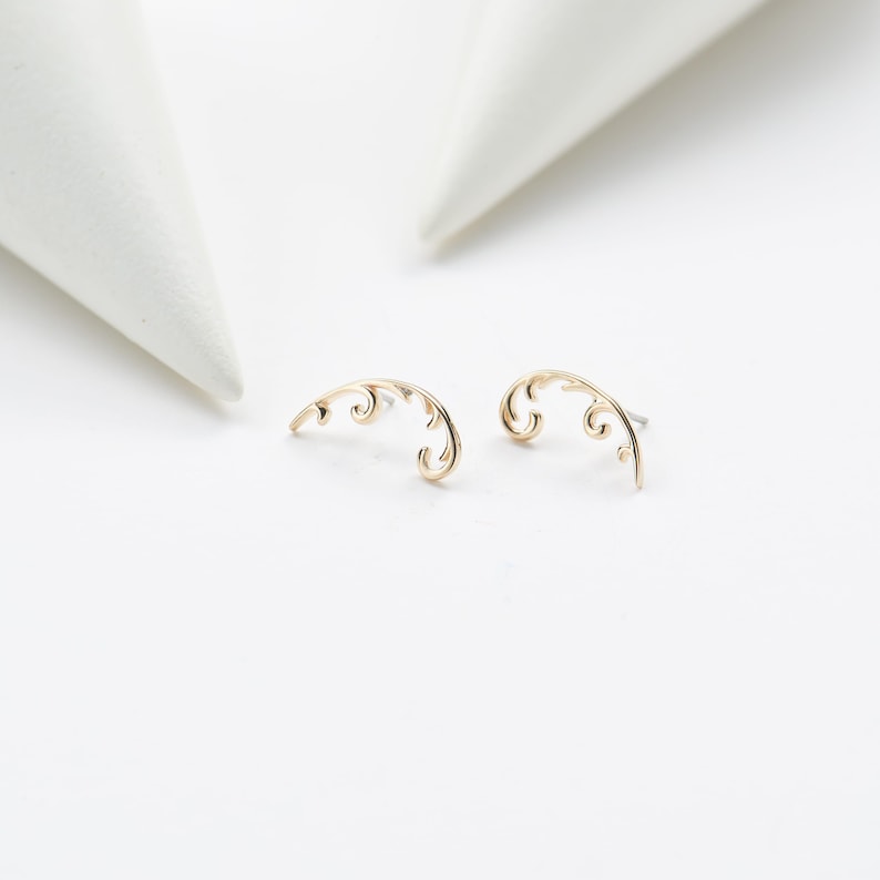 May include: A pair of gold-colored, curved earrings with an ornate, swirling design. The earrings are set against a white background, with two white, cone-shaped objects in the background. The earrings are delicate and elegant.