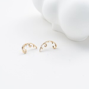May include: A pair of gold-colored, curved earrings with a delicate, swirling design. The earrings are set against a white background, with a blurred white object in the background.