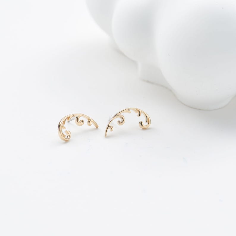 May include: A pair of gold-colored, curved earrings with a delicate, swirling design. The earrings are set against a white background, with a blurred white object in the background.