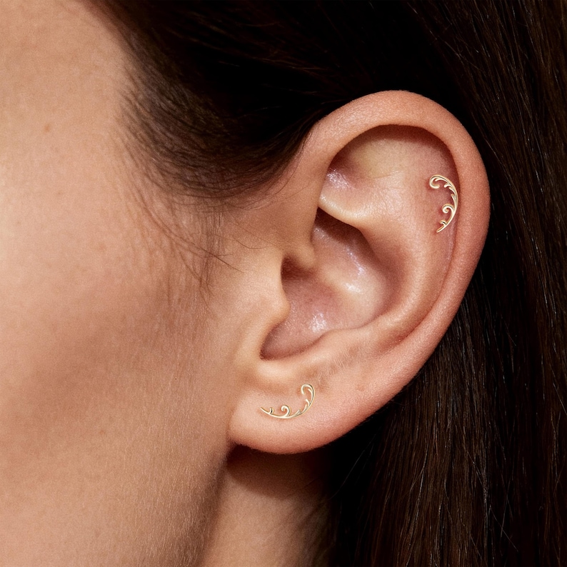 May include: Close-up of an ear adorned with two delicate, gold-colored earrings. One earring is placed on the upper ear, and the other is on the lower ear. Both earrings feature a swirling, leaf-like design, adding a touch of elegance.