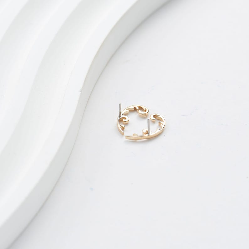 May include: A gold-colored earring with a swirling, openwork design. The earring has a post for insertion and a decorative, curved shape. The earring is set against a white background, highlighting its intricate details and elegant form.