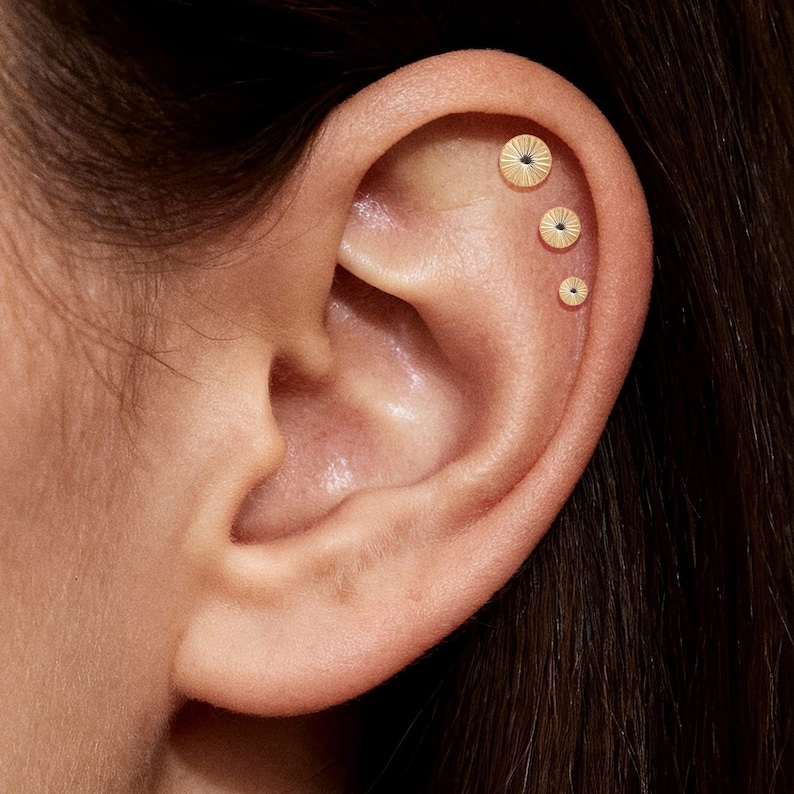 May include: Close-up of an ear with three gold-colored stud earrings. Each earring has a circular design with radial lines and a small, dark center. The earrings are of varying sizes, with the largest at the top and smallest at the bottom.