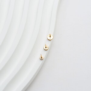 May include: Three small, gold-colored stud earrings of varying sizes are arranged on a white, wavy surface. The earrings have a circular design with a post for insertion. The background is a clean, bright white.