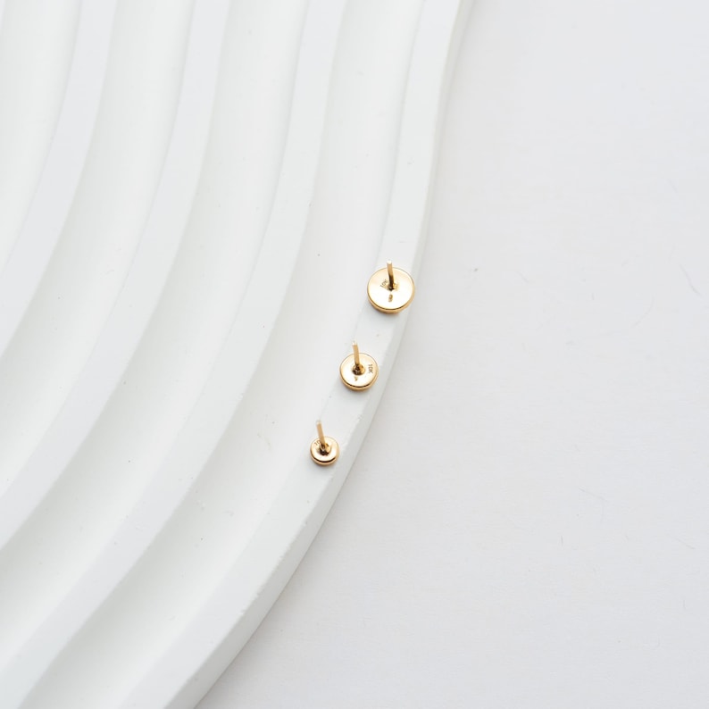 May include: Three small, gold-colored stud earrings of varying sizes are arranged on a white, wavy surface. The earrings have a circular design with a post for insertion. The background is a clean, bright white.