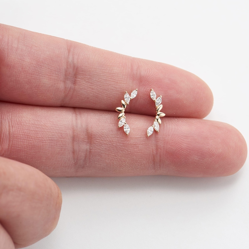 Puede incluir: Un par de pendientes dorados, cada uno con un dise&ntilde;o curvo con peque&ntilde;as piedras preciosas blancas con corte marquesa. Los pendientes tienen una apariencia delicada de hoja, con las piedras dispuestas a lo largo de la curva.