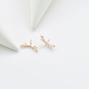 Puede incluir: Un par de trepadores de oreja dorados con un dise&ntilde;o de hojas. Cada pendiente presenta una serie de peque&ntilde;os cristales transparentes con corte marquesa. Los pendientes est&aacute;n sobre un fondo blanco, con dos formas geom&eacute;tricas blancas en el fondo.